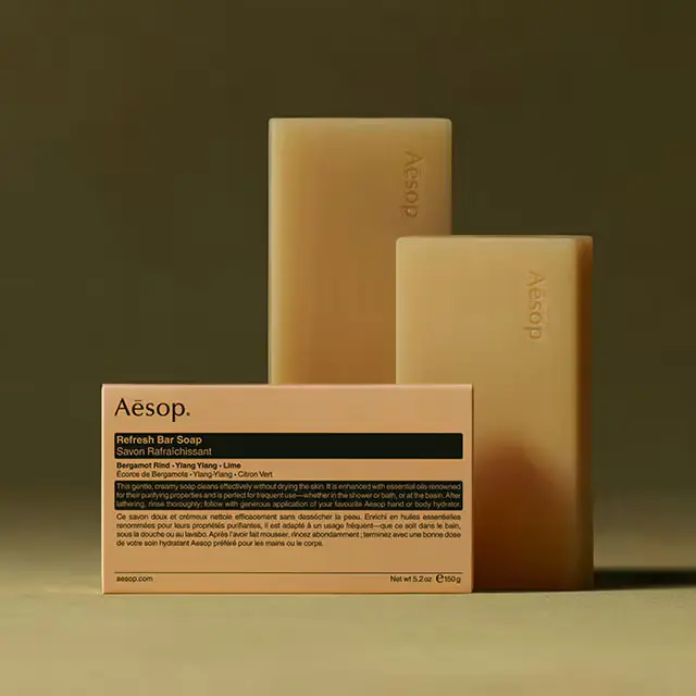 Aesop Refresh bar soaps placed horizontally behind the packaging in textured dark green background