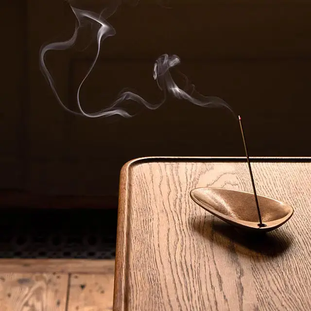 A lit incense stick resting in the Bronze Incense Holder on a wooden table.