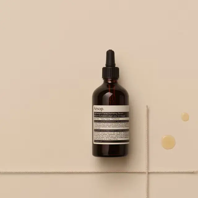 Aesop lightweight facial hydrating serum and two drops of the serum placed on textured beige background