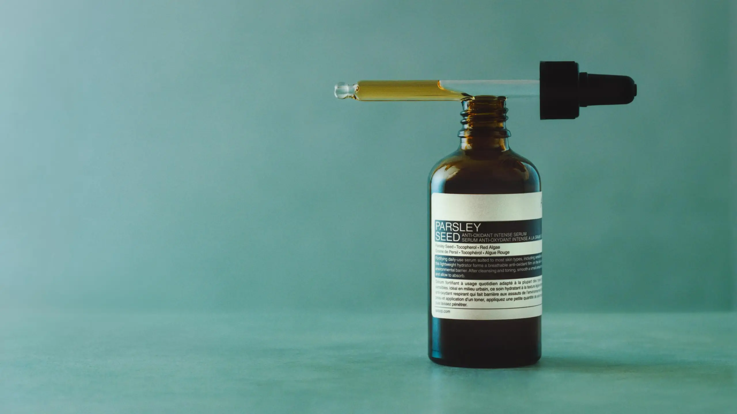 A pipette placed on top of Aesop Parsley Seed Anti-Oxidant Intense Serum in amber glass bottle, on a textured blue surface,