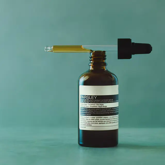 A pipette placed on top of Aesop Parsley Seed Anti-Oxidant Intense Serum in amber glass bottle, on a textured blue surface,