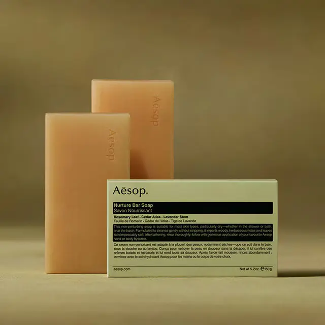 Aesop Nurture bar soaps placed horizontally behind the packaging in dark green textured background
